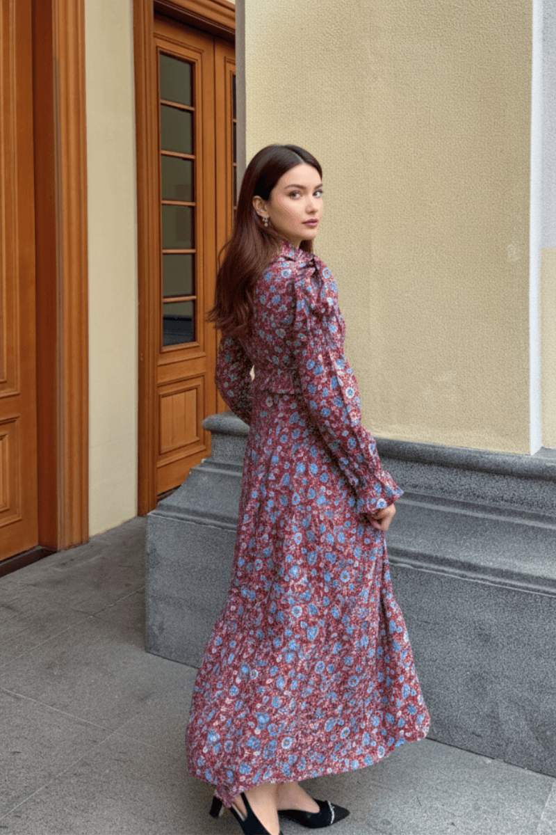 Model showing the back of a red floral print maxi dress with puff sleeves, standing outside a building with wooden doors.