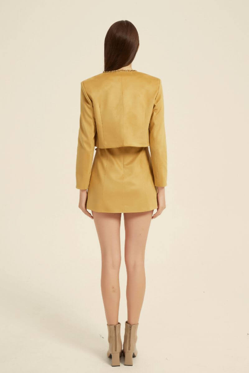 Rear view of a mustard yellow two-piece set with a structured cropped jacket and fitted mini dress, highlighting clean tailoring and soft suede-like texture.