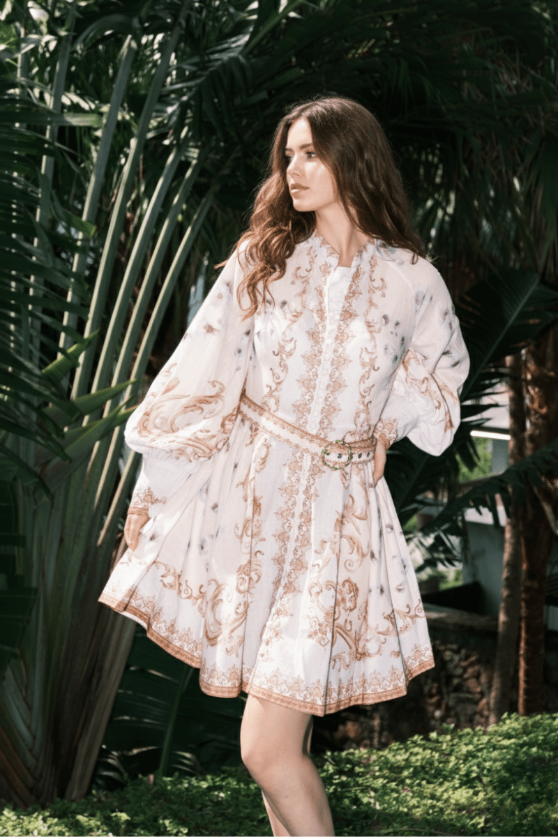 Model in a white mini dress featuring gold ornate patterns, puffed long sleeves, and a jeweled belt, standing in a lush garden with greenery background.