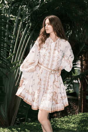 Model in a white mini dress featuring gold ornate patterns, puffed long sleeves, and a jeweled belt, standing in a lush garden with greenery background.