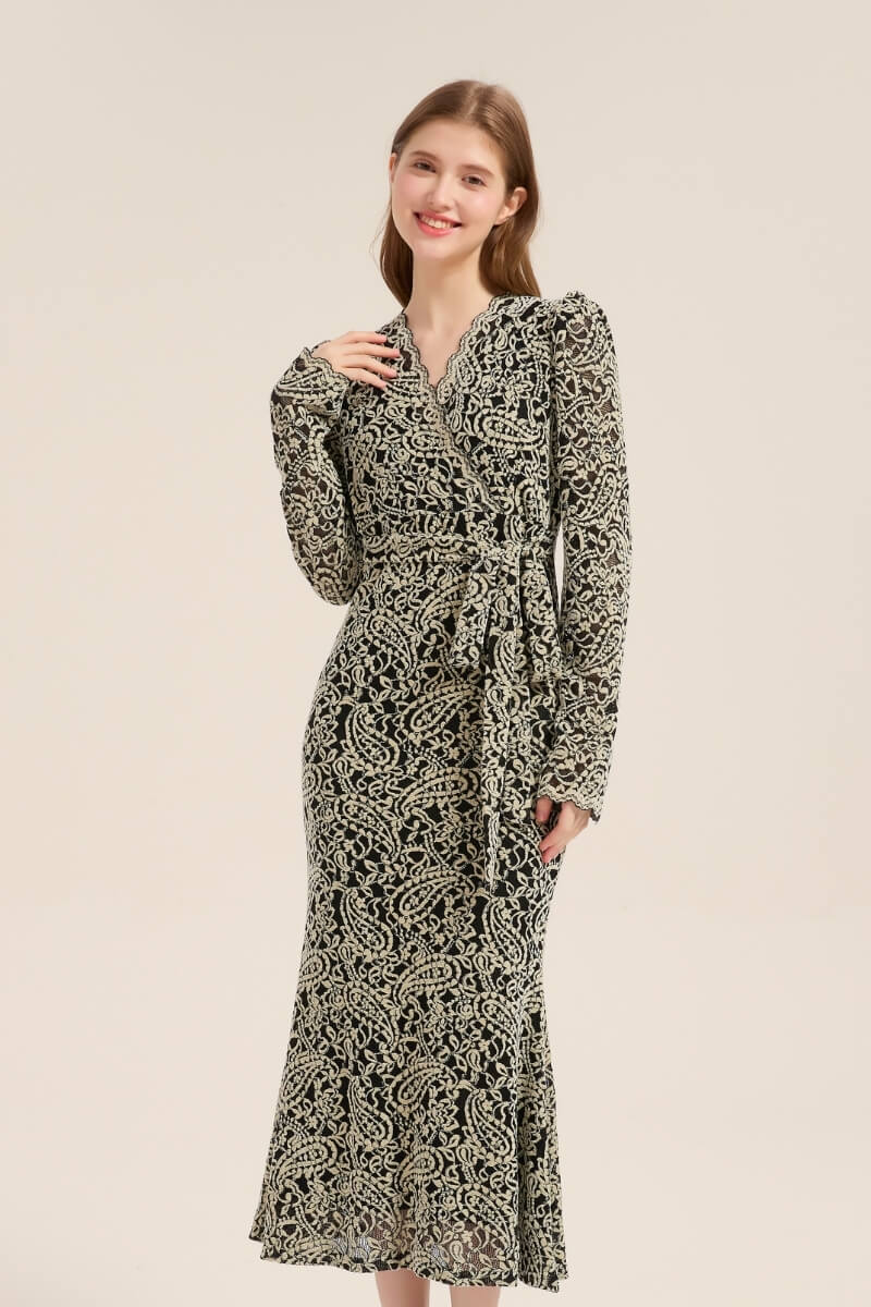 A woman wearing a black and beige floral embroidered wrap lace midi dress with long sleeves, a self-tie waist belt, and scalloped neckline, smiling softly in studio lighting.