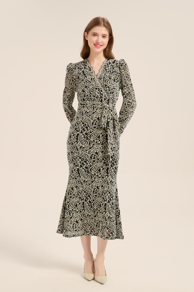 Front view of a woman in a black and beige floral lace midi dress with long sleeves, scalloped V-neckline, and a tie waist, paired with pointed heels on a beige background.