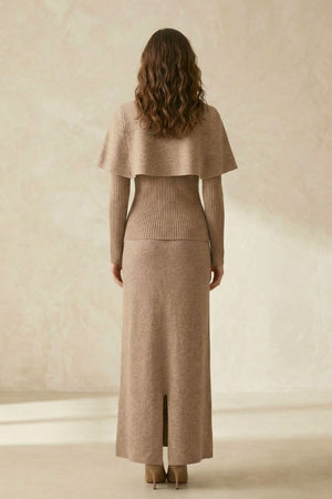 Model facing away wearing a beige knit maxi dress with a ribbed fitted waist, soft cape detail over the shoulders, and back slit, photographed in a neutral studio.