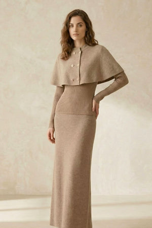 Woman wearing a beige knit maxi dress featuring a ribbed bodice, long fitted sleeves, and a cape overlay with gold-tone buttons, standing in a soft neutral-toned setting.