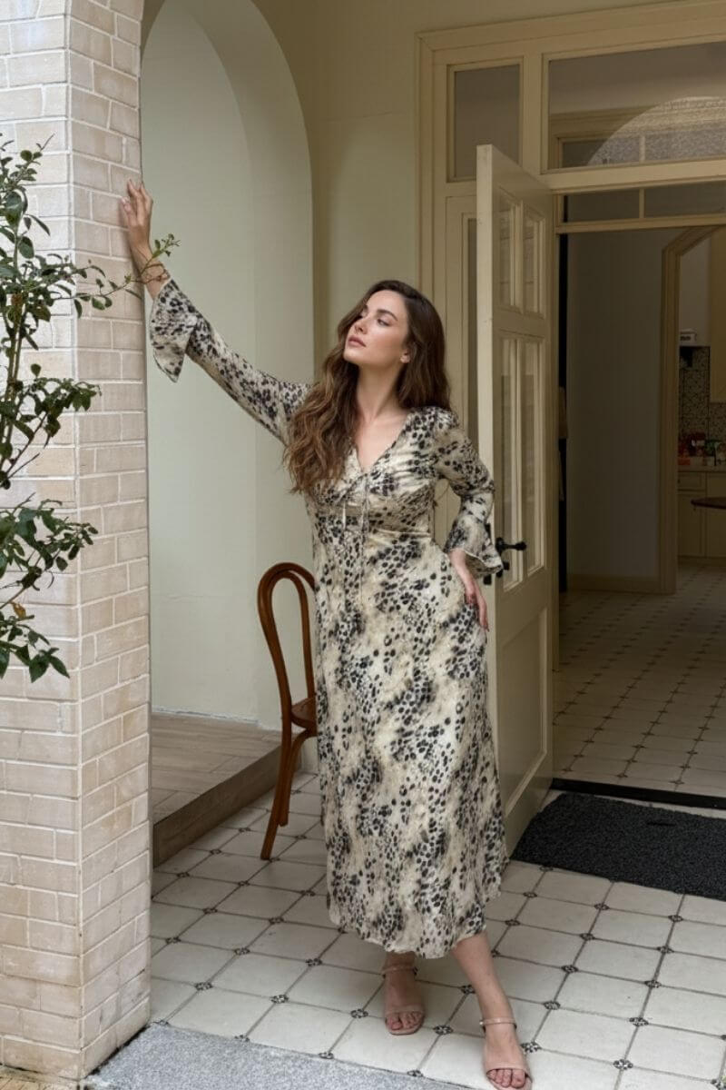 Model standing by an arched doorway wearing a beige leopard print long sleeve midi dress with a ruched V-neckline and flared cuffs, paired with nude strappy heels in a sunlit courtyard.