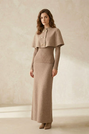 Full-body image of a model in a beige ribbed knit maxi dress paired with a structured cape overlay and gold buttons, styled with neutral ankle boots in a minimal studio.