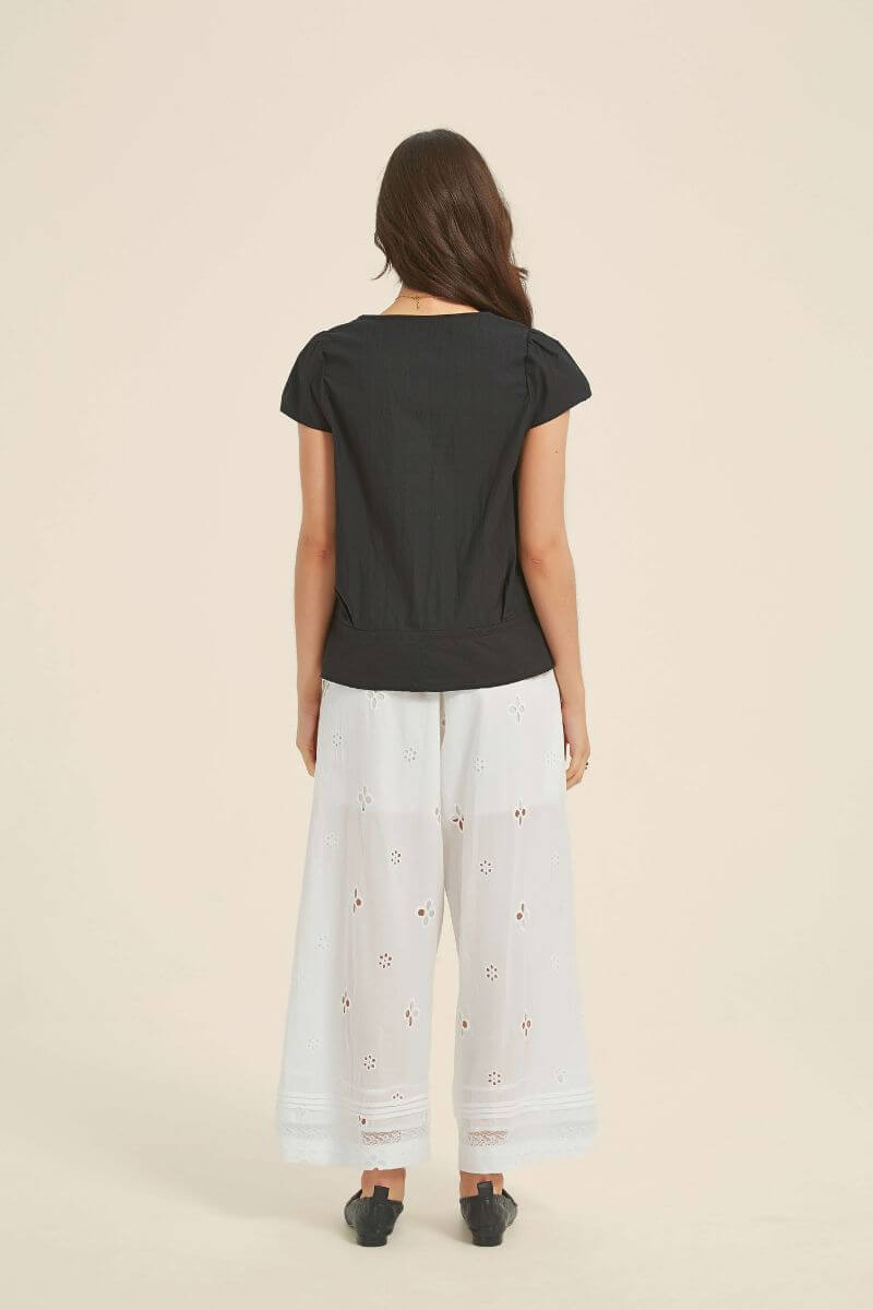 Back view of a woman in a black cotton lace-trim blouse with short sleeves and structured waist, styled with white eyelet trousers and black shoes.