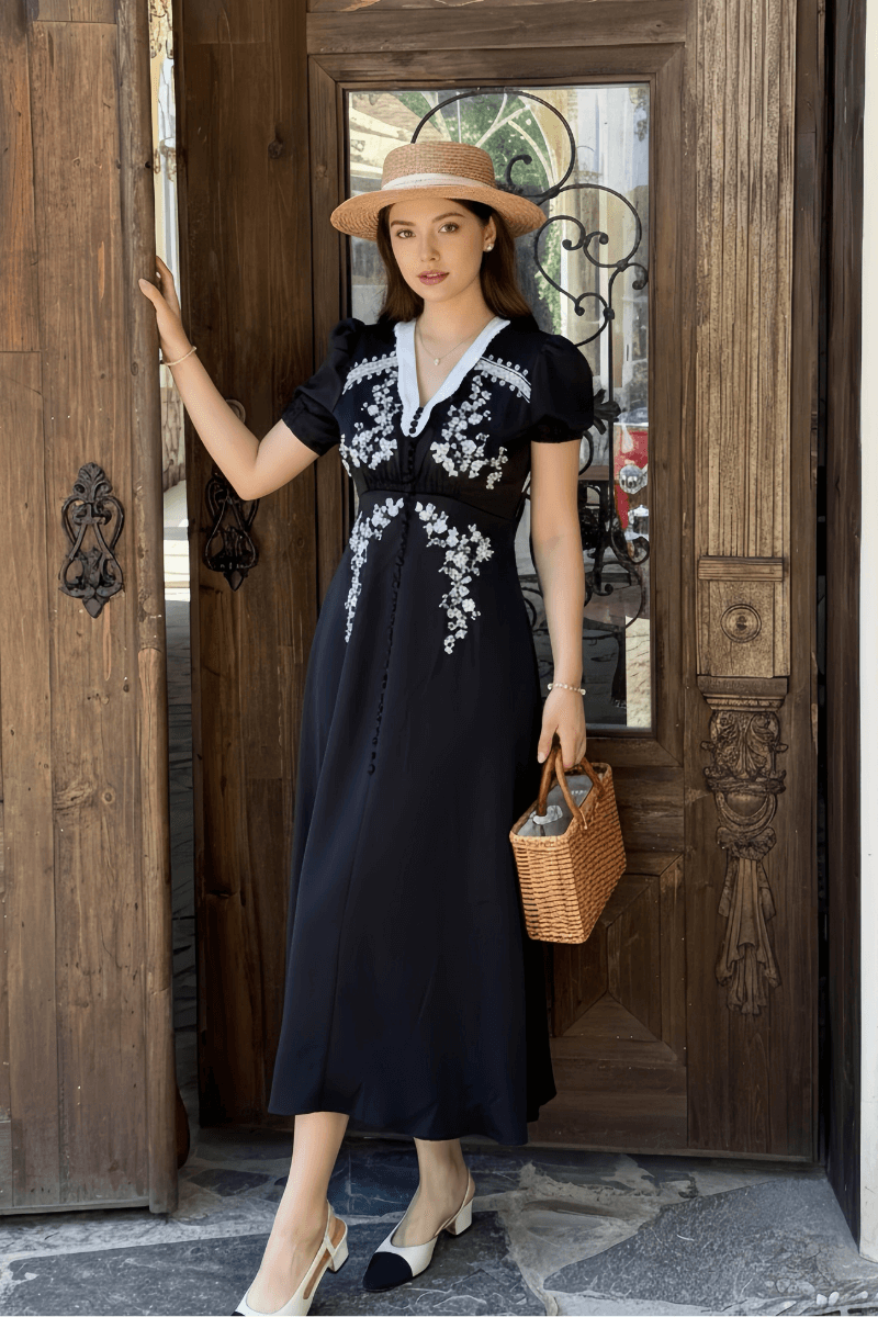A woman in a black vintage-inspired dress with white floral embroidery and a deep V neckline, accessorized with a straw hat and wicker handbag, standing in a wooden doorway.