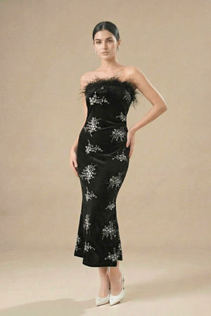 A woman in a black strapless velvet gown decorated with silver floral embellishments and a feather-trim neckline, posing elegantly in a studio.