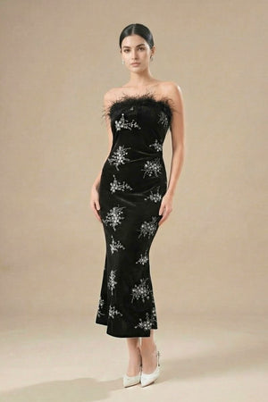 Full-body view of a woman wearing a black strapless velvet gown with feather trim and silver embellishments, paired with white heels in a studio setting.