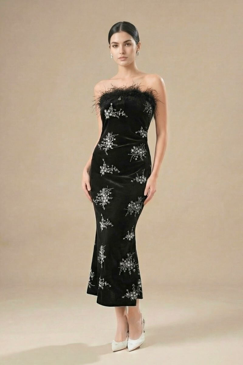 Full-body view of a woman wearing a black strapless velvet gown with feather trim and silver embellishments, paired with white heels in a studio setting.