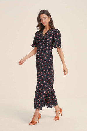 Model walking in a black floral lace-trim midi dress with flutter sleeves and subtle ruffle hem, paired with orange heels for a timeless Sioéne look.