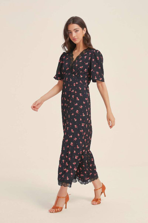 Model walking in a black floral lace-trim midi dress with flutter sleeves and subtle ruffle hem, paired with orange heels for a timeless Sioéne look.