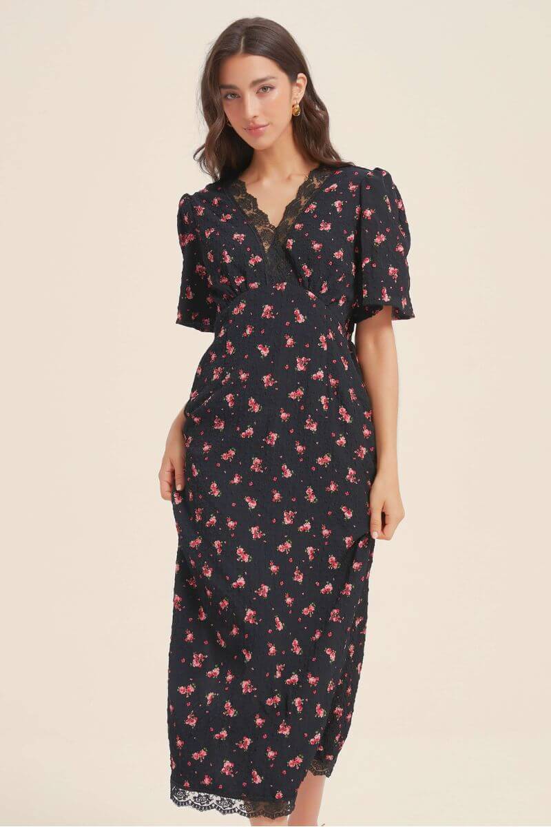 Woman wearing a black floral midi dress with puff sleeves and delicate black lace V-neck detail, featuring tiny red blossoms for a feminine vintage charm.