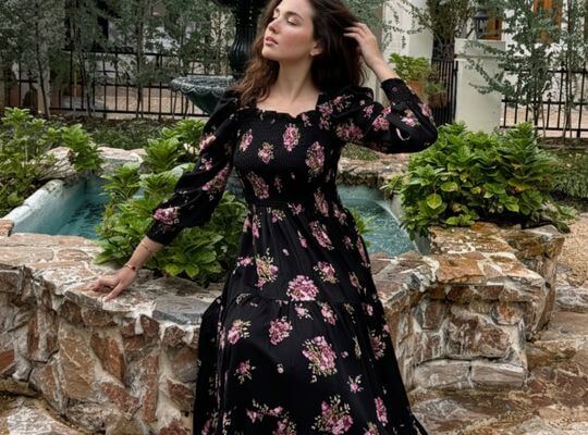 Woman in a black long-sleeve floral maxi dress sitting beside a stone fountain, styled in a soft romantic outdoor setting with pink floral patterns.