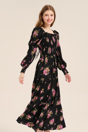 Model walking gracefully in a black floral long sleeve smocked maxi dress with tiered layers and puff sleeves, styled with white heels.