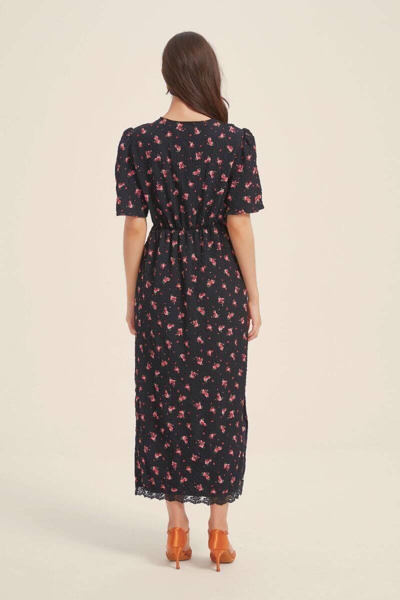 Back view of a woman wearing a black floral midi dress with an elastic waist and lace hem, styled with orange heels against a neutral background.