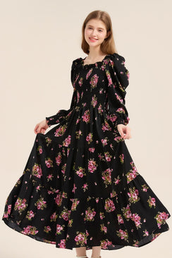 Model smiling and lifting the skirt of a black floral puff sleeve smocked maxi dress with romantic ruffle details and flowy silhouette.