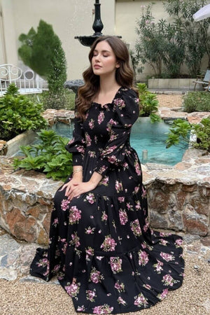 Model sitting gracefully by a stone fountain wearing a black floral smocked maxi dress with puffed sleeves and pink rose prints, eyes closed in a serene garden setting with lush greenery.