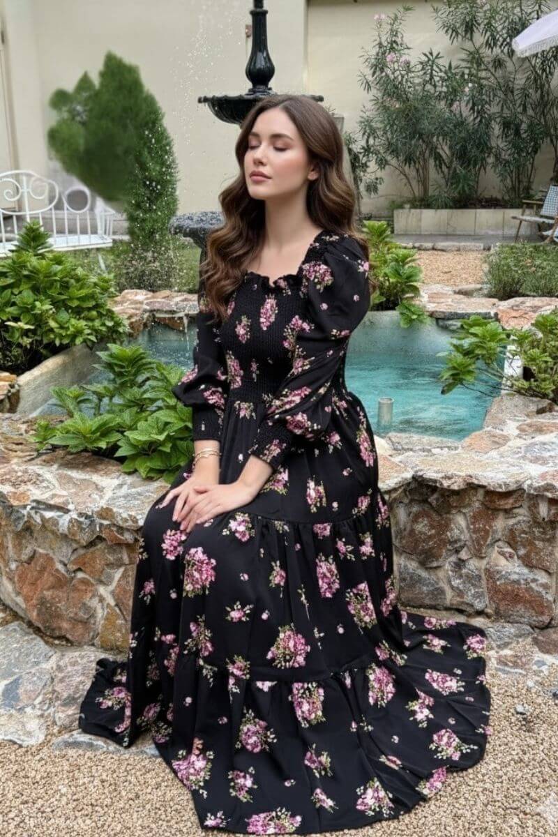 Model sitting gracefully by a stone fountain wearing a black floral smocked maxi dress with puffed sleeves and pink rose prints, eyes closed in a serene garden setting with lush greenery.