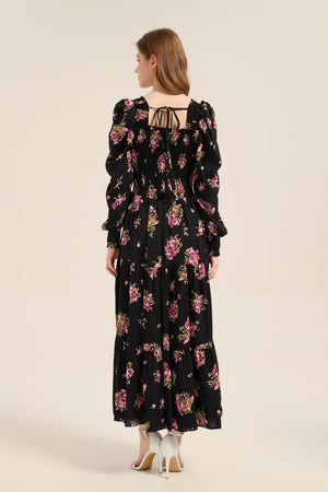 Model showing the back of a black floral smocked maxi dress featuring puff sleeves, a smocked bodice, and delicate back tie detail.