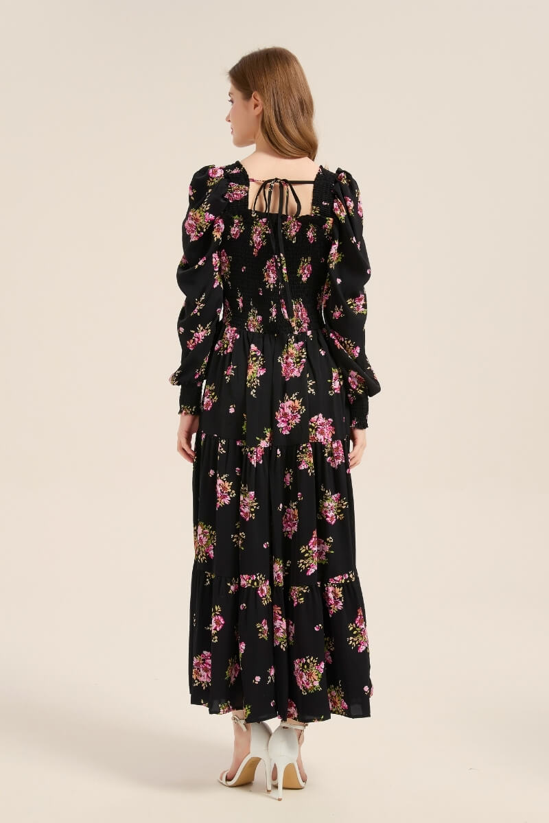 Model showing the back of a black floral smocked maxi dress featuring puff sleeves, a smocked bodice, and delicate back tie detail.