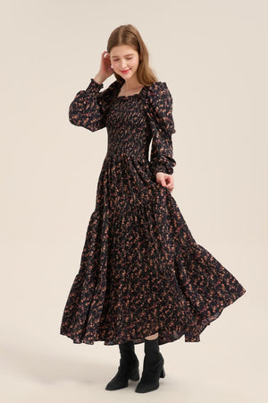 Model smiling and slightly twirling in a black floral long-sleeve smocked maxi dress with puff shoulders and a flowy tiered skirt, paired with black boots.