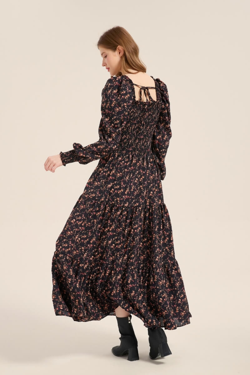 Model turning to show the back of a black floral smocked maxi dress featuring puff sleeves, tiered hem, and back tie detail, styled with black ankle boots.