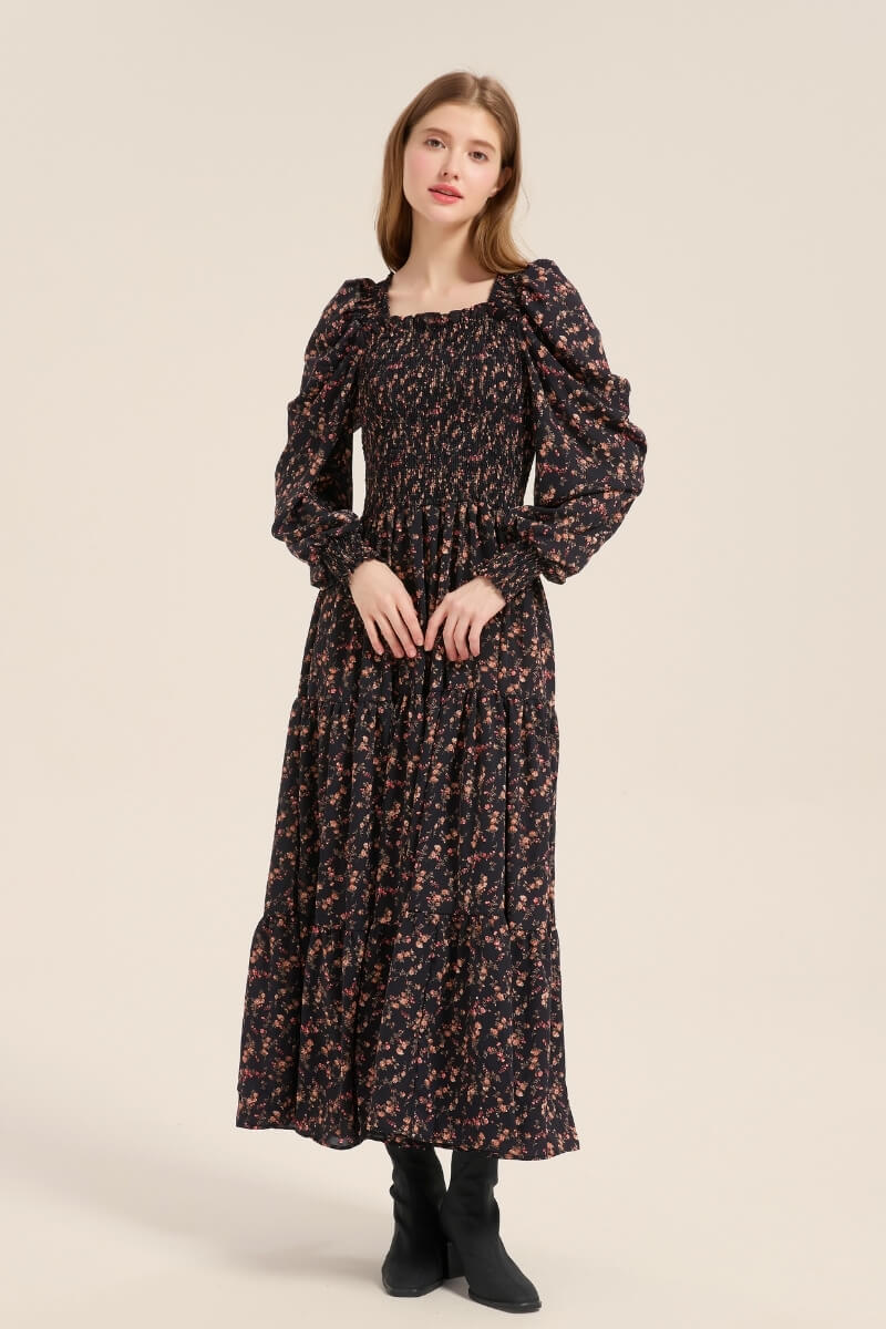 Model posing front view in a black floral smocked maxi dress with long puff sleeves and a ruffled square neckline, styled with ankle boots.