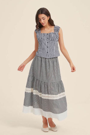 Front view of a woman wearing a black gingham two-piece outfit featuring a ruffled-strap button-front top and a tiered maxi skirt with lace trim and scalloped white hem.