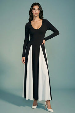 A model stands front-facing in a black long-sleeve maxi dress with a deep V neckline and flowing ivory panelled skirt, styled with white pointed heels against a soft sage backdrop.