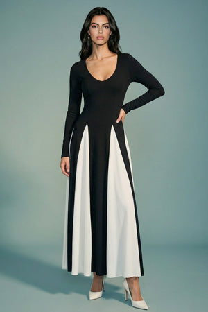 A model stands front-facing in a black long-sleeve maxi dress with a deep V neckline and flowing ivory panelled skirt, styled with white pointed heels against a soft sage backdrop.