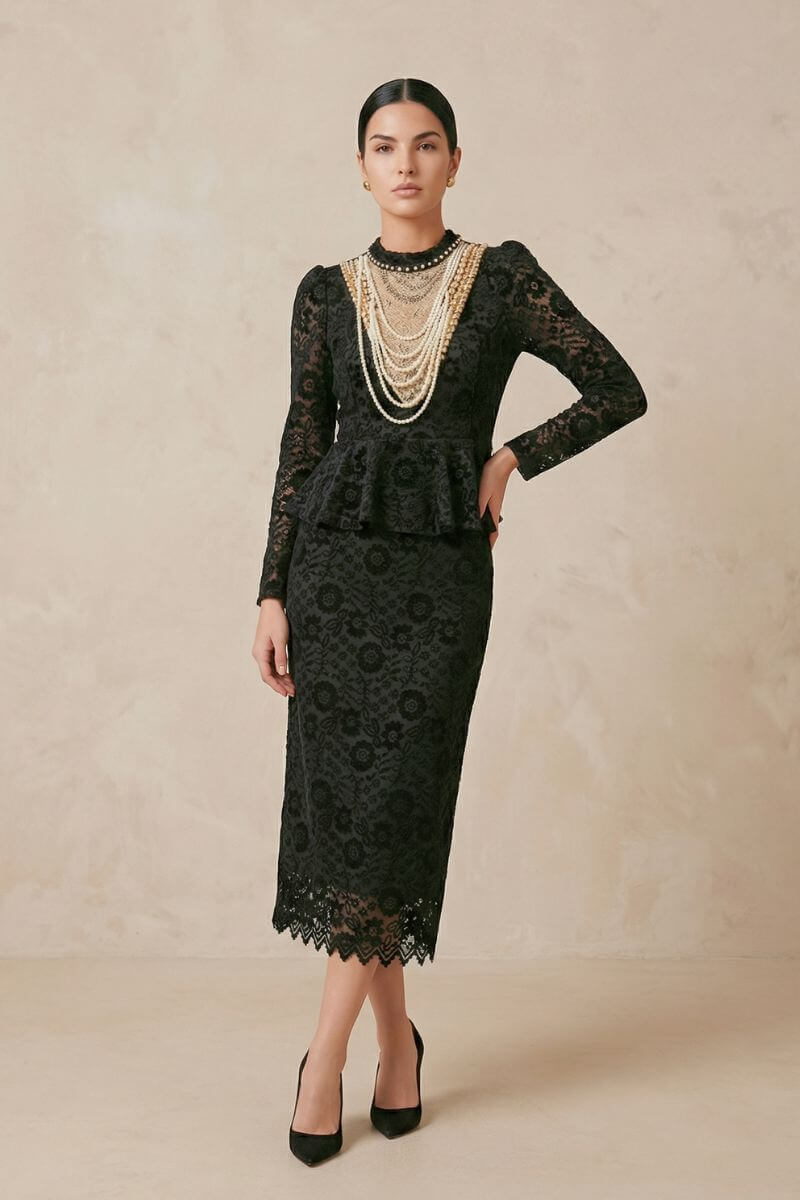 Model wearing a black lace peplum midi dress with long sheer sleeves, a high neckline layered with pearl necklaces, and a scalloped hem, styled with black heels in a warm neutral studio setting.