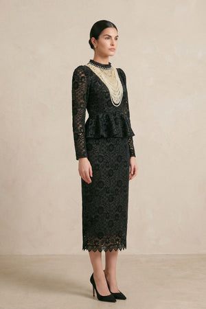 Side profile of a model in a black floral lace midi dress with long sheer sleeves, a fitted bodice, peplum waist detail, and delicate scalloped hem, styled with black heels.