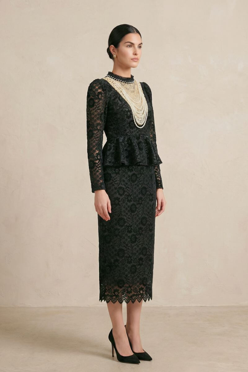 Side profile of a model in a black floral lace midi dress with long sheer sleeves, a fitted bodice, peplum waist detail, and delicate scalloped hem, styled with black heels.