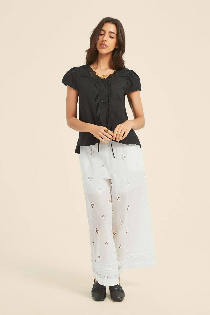 Full-length view of a woman in a black lace-trim blouse with drawstring waist, paired with white embroidered pants and black loafers for a relaxed vintage look.