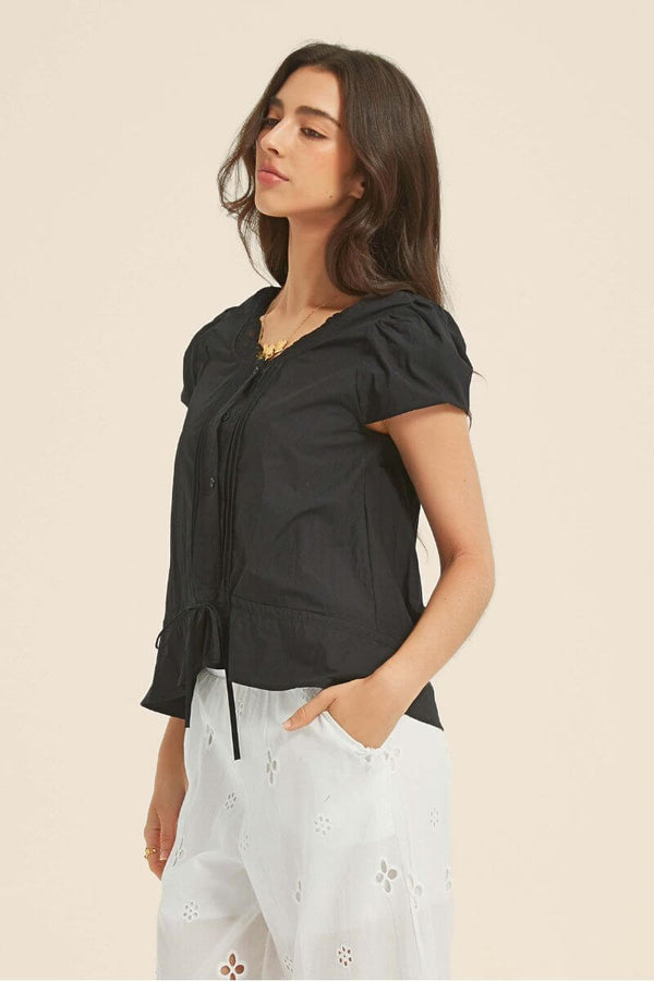 Side profile of a woman wearing a black lace-trim cotton blouse with short sleeves and tie waist, styled with white embroidered wide-leg pants.