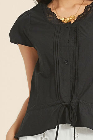 Close-up of a black blouse featuring lace neckline, front pintuck pleats, and adjustable drawstring hem, paired with white eyelet trousers.