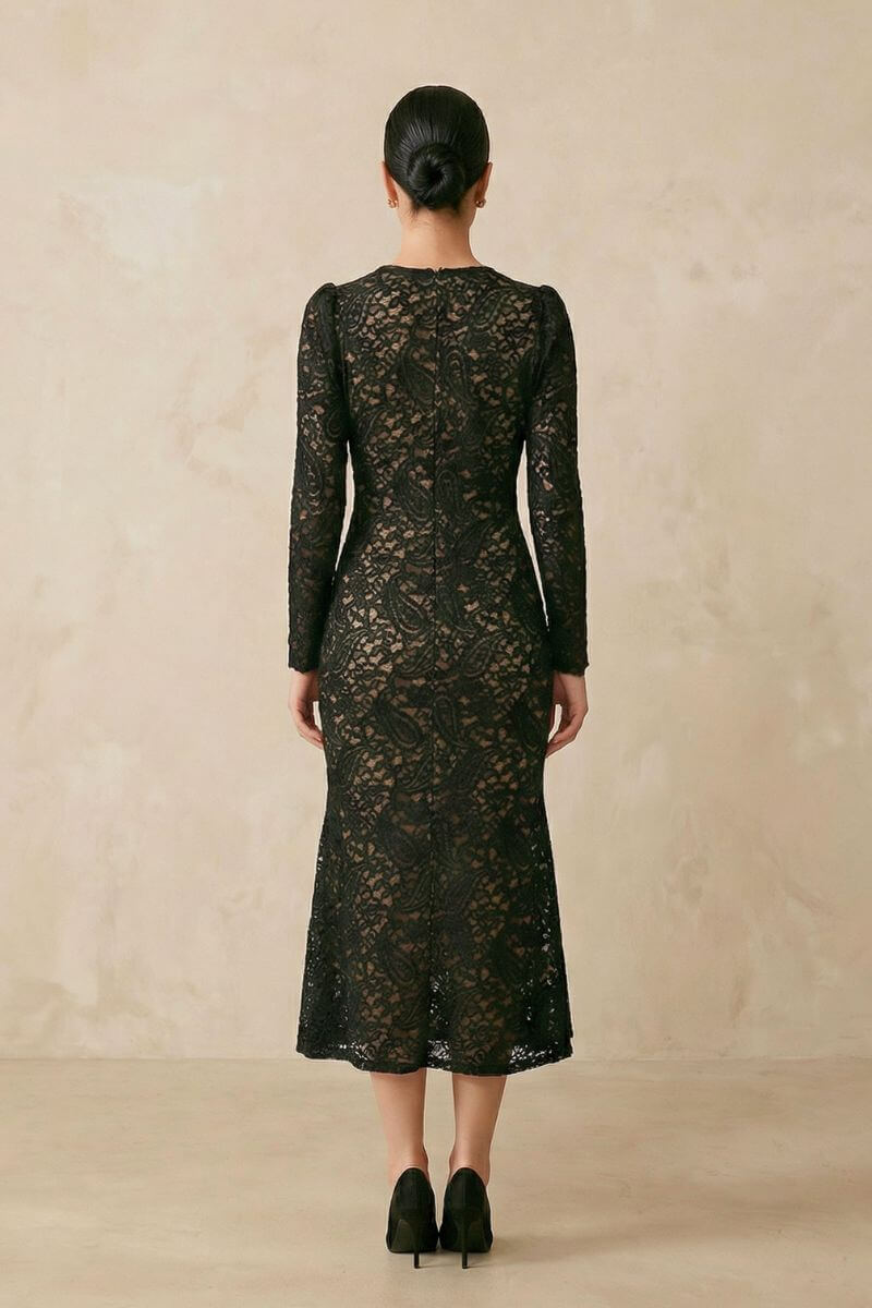 Model shown from the back wearing a black lace wrap midi dress with long sheer sleeves, a fitted waist, scalloped hemline, and a smooth low bun against a warm neutral studio background.