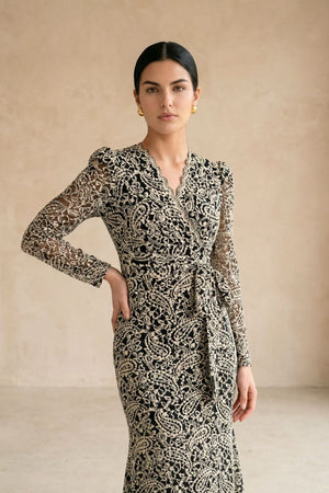 Model wearing a black lace wrap midi dress with long sheer sleeves and a softly scalloped V neckline, styled with gold earrings against a warm neutral studio background.