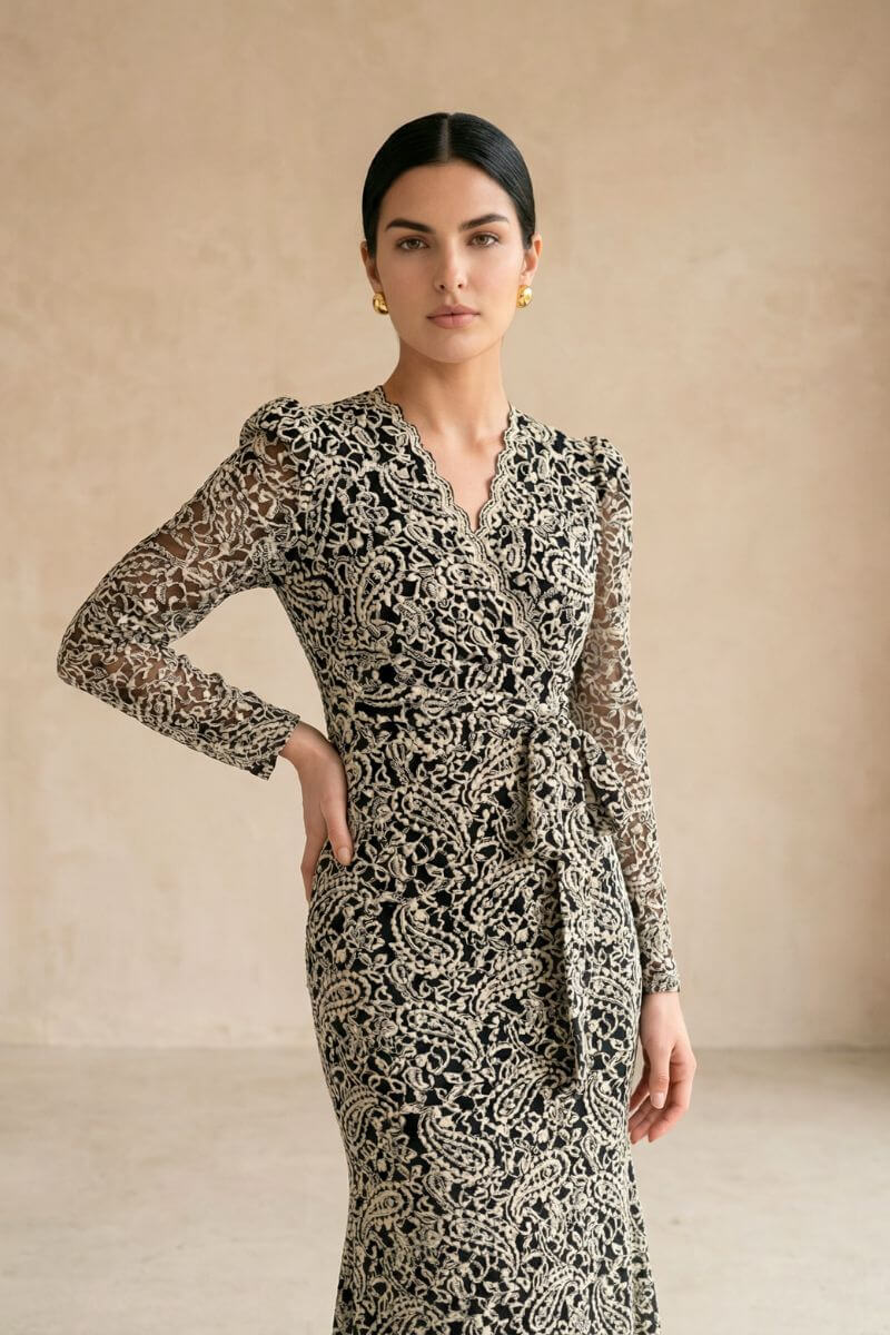 Model wearing a black lace wrap midi dress with long sheer sleeves and a softly scalloped V neckline, styled with gold earrings against a warm neutral studio background.