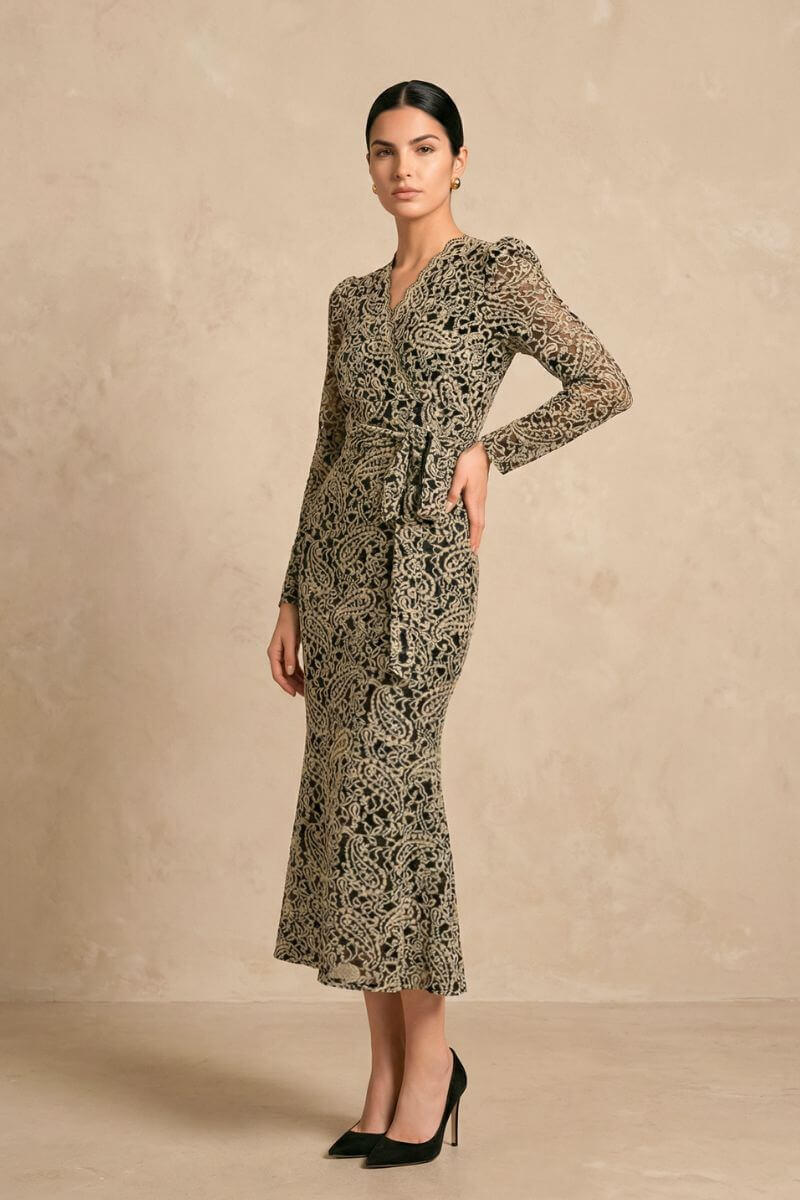 Model shown in side view wearing a black lace wrap midi dress with long sleeves and a gently defined waist tie, paired with black heels in a minimalist studio setting.