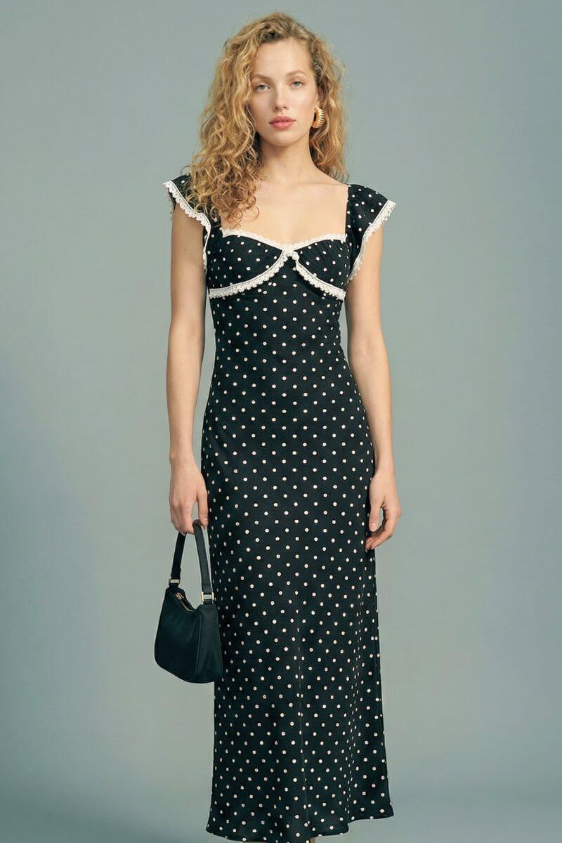 Model wears a black polka dot maxi dress with a sweetheart neckline and white lace trim, holding a black handbag against a soft gray studio backdrop.