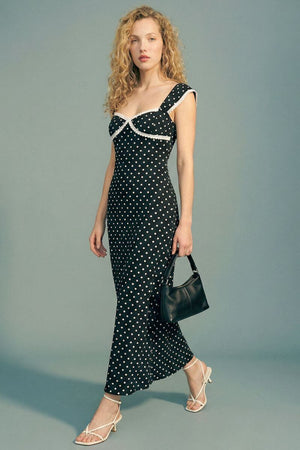 Model poses in a black polka dot lace-trim maxi dress with cap sleeves and a fitted silhouette, styled with a black handbag and white strappy heels in a gray studio setting.