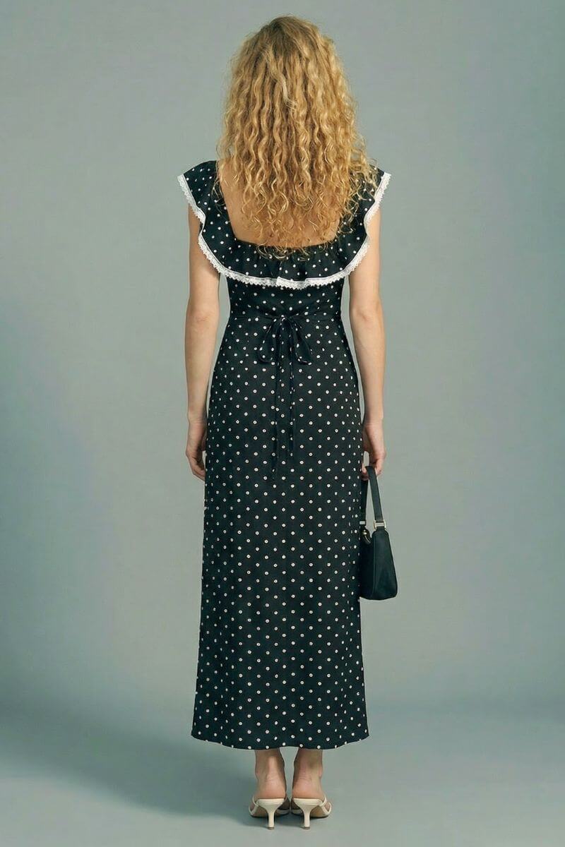 Back view of a black polka dot maxi dress with lace-trim cap sleeves and an adjustable back tie, styled with a black handbag and neutral heels on a gray backdrop.