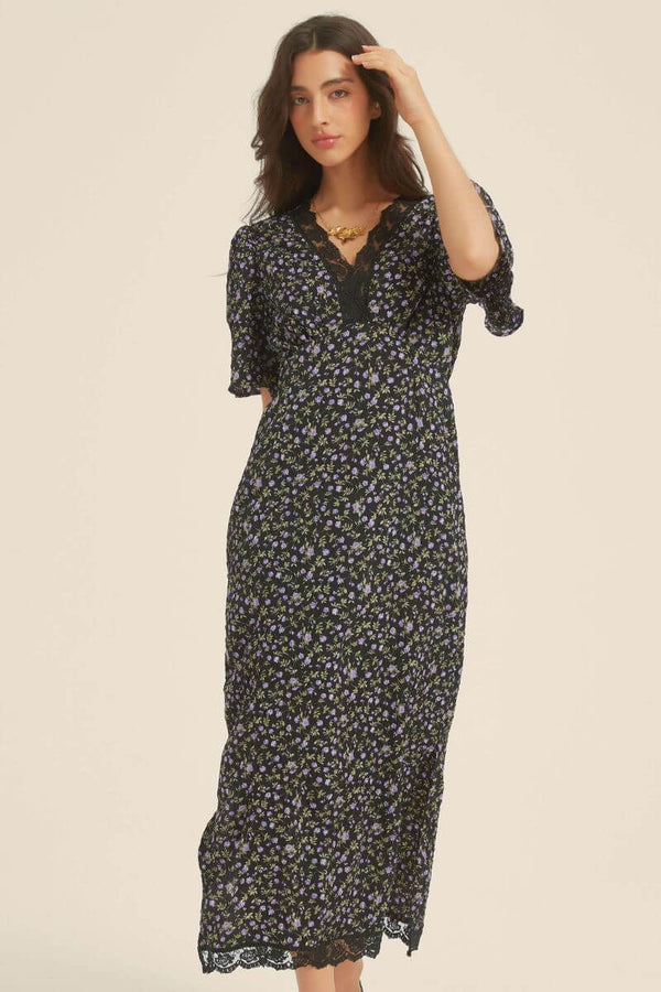 Woman in a black midi dress with purple floral print, lace-trim V-neckline, and short flutter sleeves — styled with minimal accessories for a soft romantic feel.
