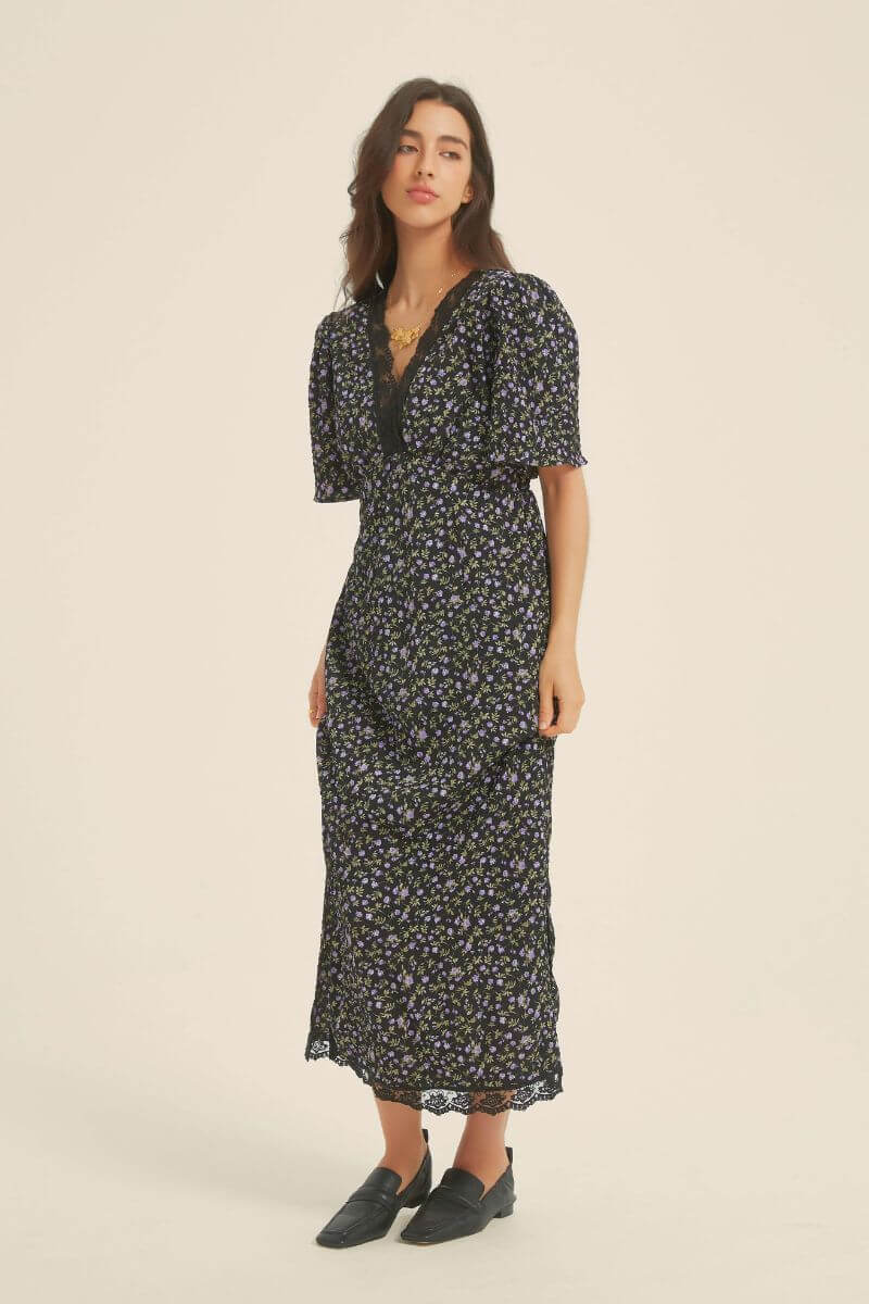Woman in a black midi dress with purple floral print, lace-trim V-neckline, and short flutter sleeves — styled with minimal accessories for a soft romantic feel.