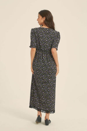 Back view of a woman wearing a black midi dress with small purple floral print, short sleeves, and lace hem — a soft romantic silhouette from Sioéne.