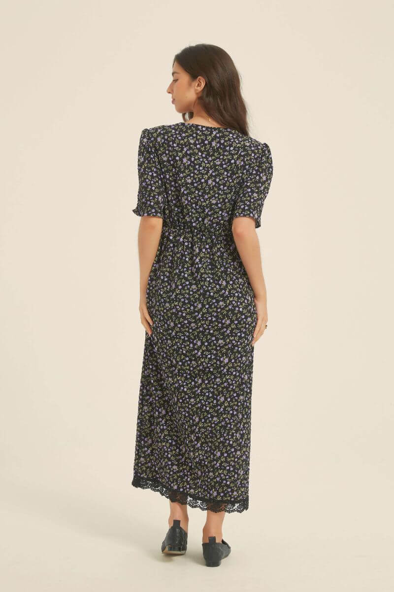 Back view of a woman wearing a black midi dress with small purple floral print, short sleeves, and lace hem — a soft romantic silhouette from Sioéne.