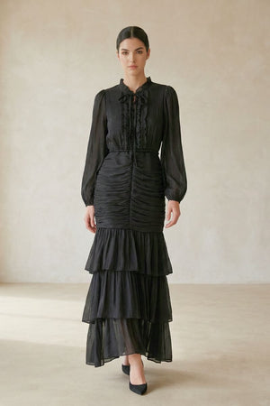 Model wearing a black ruched long-sleeve maxi dress with a tiered flowy skirt and bow-tie neckline, standing in a neutral studio setting.